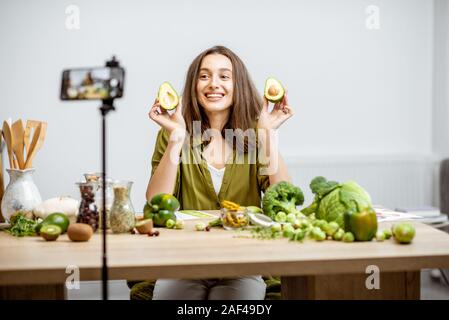 Junge Frau Aufzeichnung auf einem Smartphone Ihr vlog über gesunde Ernährung. Sitzen am Tisch mit viel Grün veganes Essen Zutaten zu Hause Stockfoto