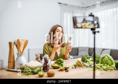 Junge Frau Aufzeichnung auf einem Smartphone Ihr vlog über gesunde Ernährung. Sitzen am Tisch mit viel Grün veganes Essen Zutaten zu Hause Stockfoto