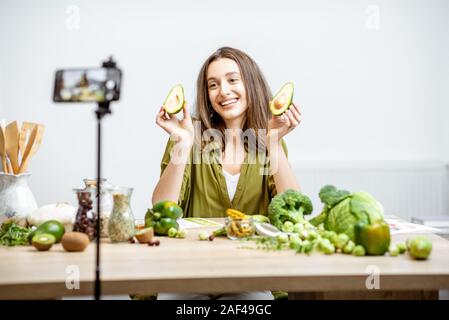 Junge Frau Aufzeichnung auf einem Smartphone Ihr vlog über gesunde Ernährung. Sitzen am Tisch mit viel Grün veganes Essen Zutaten zu Hause Stockfoto