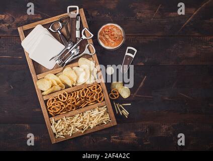 Glas Handwerk Lagerbier und Opener mit Box von Snacks auf dunklen Hintergrund. Brezeln und Chips und salzig Kartoffelstifte in vintage Holzbox mit Stockfoto