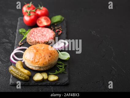 Frisches rohes Hackfleisch Pfeffer Beef Burger auf Stein Schneidbrett mit Brötchen und Zwiebeln und Tomaten auf schwarzem Hintergrund. Salzige Essiggurken und Basilikum. Ansicht von oben Stockfoto