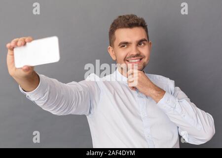 Freestyle. Junger Mann Studio stehen isoliert auf Grau unter selfie auf dem Smartphone lächelnd spielerisch zu Kamera Stockfoto