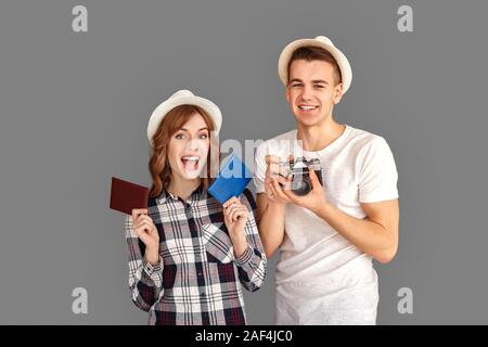 Reise und Tourismus. Junger Mann mit Kamera und Frau mit Pässen in Hüte isoliert auf Grau lächelt Fröhlich stehend Stockfoto