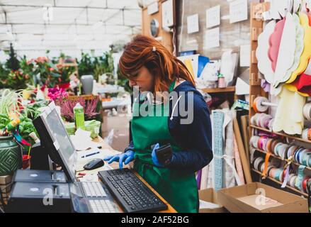 Junge Floristen mit roten Haaren arbeitet in einer Gärtnerei. Gartenarbeit Konzept. Sie arbeitet am Computer benutzt und den Barcode Scanner. Stockfoto