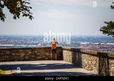 Der Mann steht mit dem Rücken auf die Aussichtsplattform in Bergamo in Italien und nimmt ein Foto auf der Kamera in Shorts, Sandalen und eine orange Stockfoto