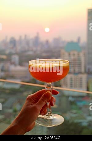 Woman's Hand mit einem Glas Cocktail mit verschwommenen Sonnenuntergang über der Skyline im Hintergrund Stockfoto