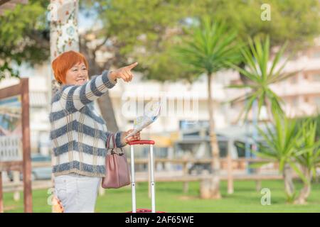 Asiatische Frau mit einer Karte und Gepäck Punkte irgendwo während sie lächelnd. Travel Concept. Stockfoto