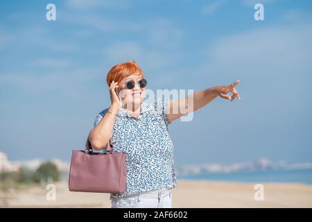 Lächelnd asiatische Frau Punkte in Richtung Strand, während Sie auf Ihrem Telefon. Urlaub Konzept. Stockfoto