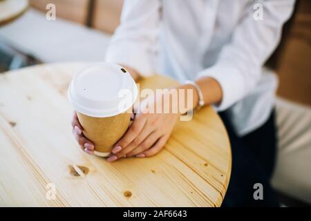 Weibliche Hände halten recyclingfähigem Papier Tasse Kaffee in einem Cafe. Ansicht von oben mit hellen Holzmöbeln. Stockfoto