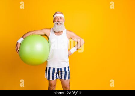 Foto von positiven Fröhliche stattlichen alten Mann hält fit Ball toothily lächelnd mit der Hand an der Hüfte selbstbewusst in der Nähe von leeren Raum lebendige isoliert Stockfoto