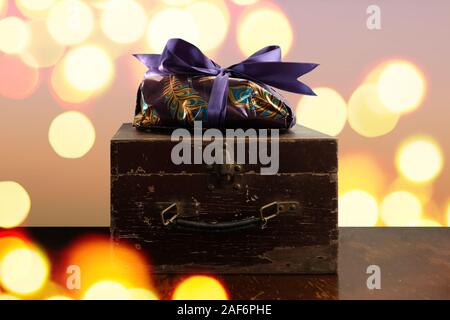 Purpur und Gold verpackt Geschenk für Weihnachten auf einer Holzkiste, gegen einen einfachen Hintergrund mit funkelnden Lichtern. Stockfoto