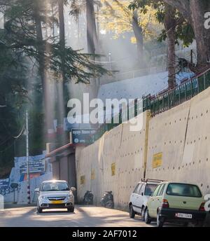 Lichtschächte filtern in den frühen Morgenstunden in der Himalaya-Stadt Almora durch die Bäume. Ein Auto von Maruti Suzuki fährt die Straße hinunter. Stockfoto