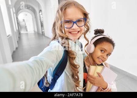 Zwei Mädchen blond Kaukasischen mit Locken und afrikanischen Mädchen tun selfie Korridor in der Schule zusammen. Schöne und lustige Freunde mit Noten und Kopfhörer Stockfoto