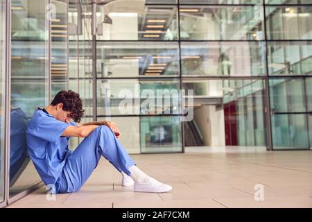 Müde Junge männliche Arzt tragen Scheuert sich gegen Wand im modernen Krankenhaus Gebäude Stockfoto