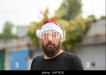 Santa Claus zu Fuß auf der Straße der Stadt. Mann mit Bart auf der Straße im Sommer in einen Hut von Santa Claus. Ein Mann mit einem emotionalen Bart stellt ein Bac Stockfoto