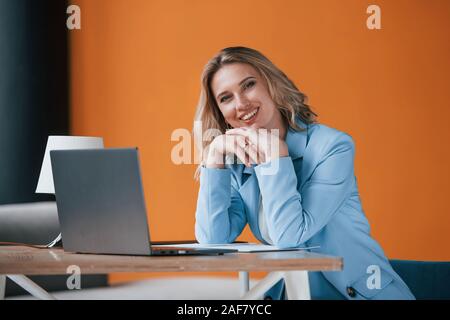 Immer glücklich, Sie zu sehen. Geschäftsfrau mit lockigem Haar drinnen im Zimmer mit orange farbigen Wand und Holztisch Stockfoto