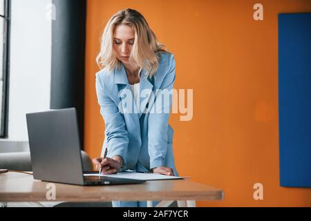 Lassen Sie die Arbeit beginnen. Geschäftsfrau mit lockigem Haar drinnen im Zimmer mit orange farbigen Wand und Holztisch Stockfoto