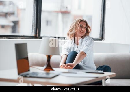 Mit dem neuen Büro zufrieden. Geschäftsfrau mit Lockigen, blonden Haaren sitzen im Zimmer gegen Fenster Stockfoto