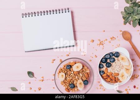Gesundes Müsli Frühstück mit Muttern und Heidelbeere neben leer Notebook auf rosa Tabelle, flach. Stockfoto