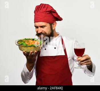 Koch hält Schüssel mit Kartoffel Auflauf und Glas Alkohol. Kochen mit zufriedenen Gesicht in Burgund einheitliche riecht gebackene Speise und Wein. Mann mit Bart auf weißem Hintergrund. Sommelier und kochen Konzept Stockfoto