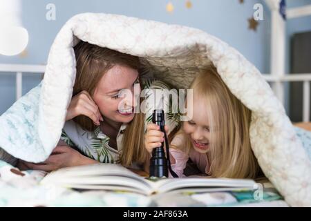 Junge Mutter lachend mit glücklichen Tochter mit einem Buch unter der Bettdecke mit Taschenlampe Stockfoto