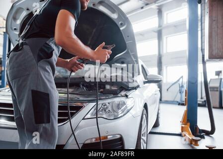 Hält rote Überbrückungskabel. Mann in der grauen uniform Reparaturen weiß Automobil im Innenbereich Stockfoto