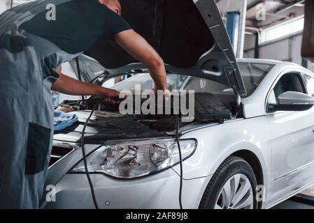 Mit Überbrückungskabel. Mann in der grauen uniform Reparaturen weiß Automobil im Innenbereich Stockfoto