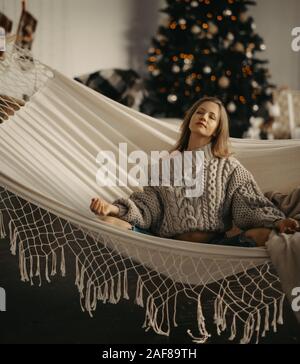 Junge schwangere Frau in Pullover sitzt und meditiert in Yoga Pose in der Hängematte vor Hintergrund der Weihnachtsbaum. Stockfoto