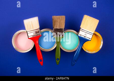 Vier offene Farbdosen mit Bürsten auf blauem Hintergrund. Gelb, Blau, Pink, Türkis Farben. Ansicht von oben. Stockfoto