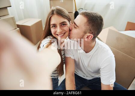 Machen Sie ein Foto. Kerl küsst seine Frau. Fröhlicher junger Paare in der neuen Wohnung. Konzeption von verschieben Stockfoto