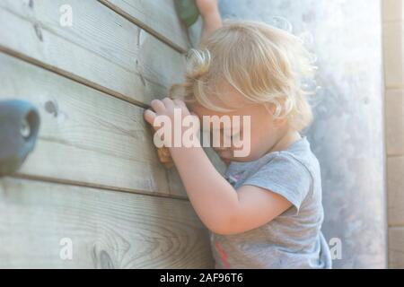 Portrait von cute blonde Mädchen klettern Kunststoff Boulderwand outdoor. Stockfoto