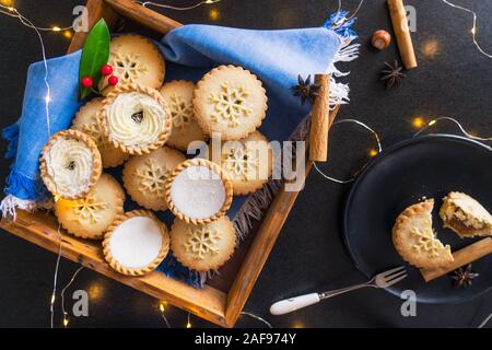 Ansicht von oben warmen gemütlichen Zusammensetzung des traditionellen englischen festliches Gebäck Mince Pies in Holz- fach, Gewürze, schwarze Platte mit gebrochenen Pie und Lichter Girlande Stockfoto