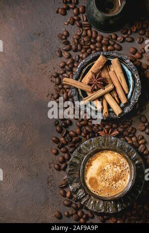 Schwarzen Kaffee Espresso mit Schaumstoff in schwarz Keramiktasse mit Untertasse, cezve Kaffeekanne, Gewürze und gerösteten Bohnen über braune Textur Hintergrund. Fla Stockfoto
