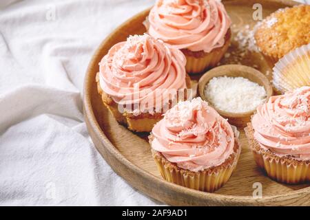 Hausgemachte Muffins mit Rosa buttercream und Kokosraspeln in Holz- Platte über weiße gefalteten Tischdecke serviert als Hintergrund. Nahaufnahme Stockfoto