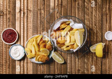 Klassische britische fast food Fisch und Chips in den Frittierkorb mit Zitronen serviert, roter und weißer Soße, Salz in Keramik Schalen über holzbrett Hintergrund. Stockfoto