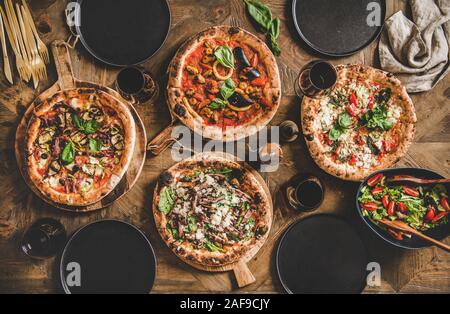 Pizza Party Abendessen. Flachbild-lay von verschiedenen Arten von italienischer Pizza, frischem Salat und Rotwein Gläser über rustikalen Holztisch, Ansicht von oben. Fast food lunc Stockfoto