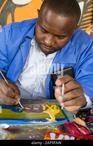 Arusha, Tansania. Künstler Malerei im Tinga-Tinga Stil in Shanga, einem Zentrum für Kunsthandwerk Beschäftigung von Behinderten. Stockfoto