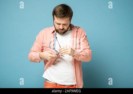 Missfallen bärtiger Mann seine Brille reinigt von Staub und Schmutz, wischen Sie mit einem T-Shirt. Auf einem blauen Hintergrund. Stockfoto