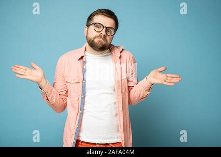 Bärtiger Mann mit einem unschuldigen Blick zuckt, breitet die Arme in die Seiten und sieht in die Kamera. Auf einem blauen Hintergrund. Stockfoto