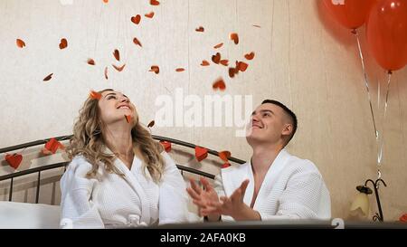 Liebevolle Freundin und Freund in bademänteln haben Spaß im Bett. Sie werfen Konfetti von roten Herzen. Stockfoto