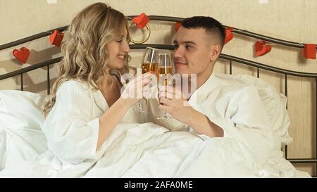 Junges Paar in Liebe im Bett klirren Gläser mit Champagner. Romantischer Abend im Zimmer Stockfoto