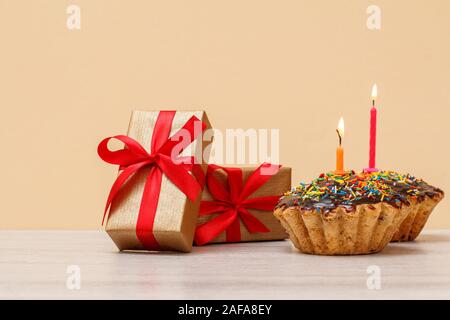 Lecker geburtstag Muffin mit Schokoladenüberzug und Karamell, mit brennenden festliche Kerze und Geschenkboxen mit roten Bändern auf dem beigen Hintergrund eingerichtet. Stockfoto