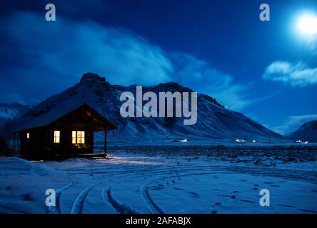 Landschaft mit einem Holzhaus mit Licht vom Fenster in einer Winternacht. Malerischer Blick auf Mondlicht auf dem Schnee mit Bergen im Hintergrund. So Stockfoto