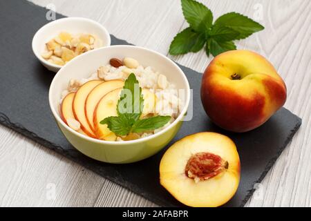 Porzellan Schüssel mit sorghum Porridge mit Cashew Nüssen und Mandeln, ganz, frisch peache und eine Hälfte eine kleine Schüssel mit Muttern auf Stein. Vegan glutenfrei Stockfoto