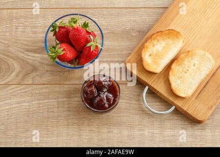 Hausgemachte Erdbeermarmelade in eine Glasschüssel, frische Beeren, Toast auf Schneidebrett auf hölzernen Schreibtisch. Ansicht von oben. Stockfoto