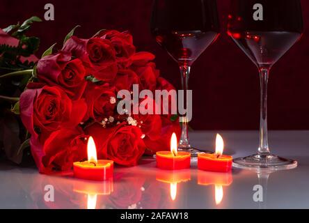 Blumenstrauß aus roten Rosen und zwei Gläser Wein mit brennenden Kerzen auf eine reflektierende weiße Tischplatte Stockfoto