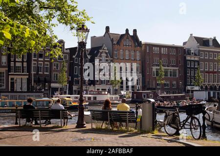 Anzeigen von Menschen Standortwahl auf Bänken betrachten Menschen Reiten eine Tour Boot am Fluss Amstel in Amsterdam. Traditionelle Straßenlaterne und historische Bauten Stockfoto