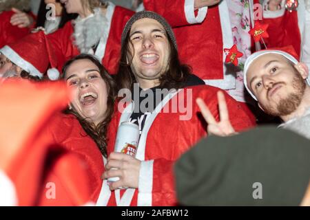Trafalgar Square, London, Großbritannien. Dezember 2019. Die Teilnehmer des Santacon 2019 treffen sich auf dem Trafalgar Square, London, in festlichen Weihnachtsmannskostümen während einer nächtlichen Feier. Das Bild fängt eine fröhliche Szene mit Einzelpersonen ein, die posieren, lachen und sich in Feiertagsstimmung einmischen. Die zentrale Figur hält eine Dose Stella Artois, während andere rote Mäntel und weiße Mützen tragen und Friedenszeichen machen. Der beleuchtete Platz und die umliegende Architektur bilden eine dramatische urbane Kulisse für dieses verspielte saisonale Treffen. Penelope Barritt/Alamy Live News Stockfoto