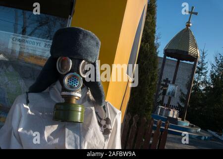 Ein ausgestellt Dummy tragen ein Schutzanzug und Gasmaske außerhalb eines Shop mit Souvenirs am Eingang der Sperrzone von Tschernobyl am Checkpoint "ytyatky'. Im Hintergrund: Eine kirchliche Denkmäler für die Victums der Katastrophe von Tschernobyl im Jahr 1986. Sperrzone von Tschernobyl, Checkpoint, Dytyatky Invankiv Rajon, Kiev Oblast, Ukraine Stockfoto