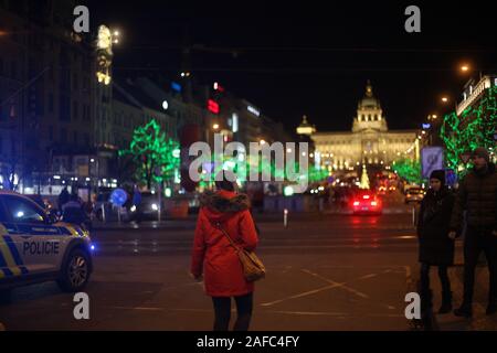 Das Leben auf der Straße - Rückansicht einer Frau in einem roten Mantel & Menschen zu Fuß über eine Straße Kreuzung bei Nacht. Das Zentrum Prags. Der Tschechischen Republik. Dezember 2019. Stockfoto